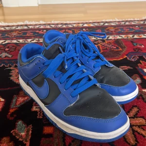Nike Dunk Low blå och svarta sneakers - Nike Dunk Low sneakers i blått och svart med vit sula. Skorna har klassisk snörning, rund tå och platt sula. Ovandelen är tillverkad i skinn och syntet, med blå Nike-logga på sidan. Perfekt för dig som gillar streetwear och sportig stil.