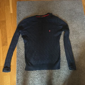 Mörkblå kabelstickad tröja Morris XS - Säljer en mörkblå kabelstickad tröja från Morris i storlek XS. Tröjan har lång ärm, rund hals och en rosa broderad logga på bröstet. Klassisk och stilren modell med ribbade muddar och snygg struktur i materialet.