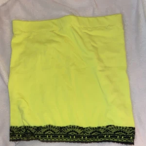 Neon grön/gul kortkjol med svart spetskant - Snygg gul kortkjol med svart spetsdetalj längst ner. Kjolen har en rak och enkel form, perfekt för dig som gillar färgstarka plagg med en twist. Den är tillverkad i mjukt bomullsmaterial och har en elastisk midja för skön passform.