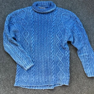 Blå kabelstickad polotröja Clifton Classic - Kabelstickad blå polotröja från Clifton Classic i 100% bomull. Tröjan har ett klassiskt flätmönster, hög krage och långa ärmar. Perfekt för dig som gillar en tidlös och chill look med lite extra struktur.