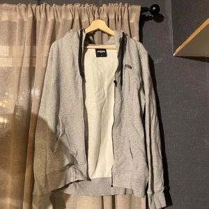 Grå hoodie från Pearson med dragkedja - Grå hoodie från Pearson med svart logga på bröstet. Tröjan har hel dragkedja, huva med svarta snören och två fickor framtill. Insidan är mjuk och fleecefodrad, perfekt för kyliga dagar. Klassisk och enkel stil som funkar till det mesta.