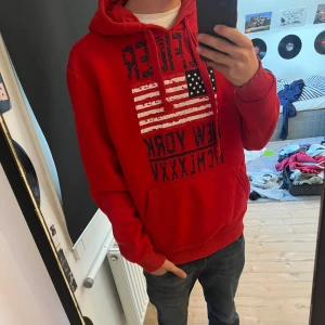 Röd hoodie med USA-flagga och tryck - Röd hoodie från Tommy Hilfiger med stor amerikansk flagga och texten 'New York' på bröstet. Tröjan har huva med snörning, magficka och långa ärmar. Perfekt för dig som gillar streetstyle och vill sticka ut med en färgstark look.