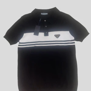 Svart och vit Prada piké - Snygg svart pikétröja från Prada med vit panel och dubbla ränder över bröstet. Klassisk krage, korta ärmar och knappslå framtill. Ikonisk Prada-logga på bröstet ger en exklusiv touch. Perfekt för dig som gillar stilrena och sportiga vibes.