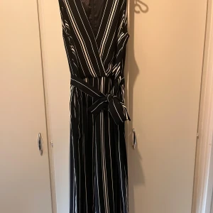 Jumpsuit strl 40 - Randig jumpsuit i svart och vit. Strl 40.  Från H&M.  Byxbenen är vida, trekvart längd.  Knytning i midja och fickor fram.  Dold dragkedja i ryggen.  Fint skick, se bild nr 4, då det är ett litet märke i tyget baktill men inget man tänker på. Material: polyester 