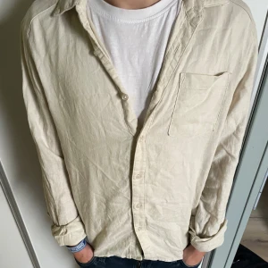 Beige linneskjorta med bröstficka - Snygg beige skjorta i linne från ryvls med klassisk krage och bröstficka. Skjortan har långa ärmar och knäppning framtill. Perfekt för en avslappnad och stilren look.