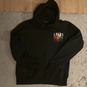 Svart hoodie från Amiri med regnbågstryck - Svart hoodie från Amiri med färgstarkt regnbågstryck och drip-effekt både fram och bak. Stor huva, klassisk känguruficka och coola logodetaljer. Perfekt statement-plagg för dig som gillar streetwear och vill sticka ut.