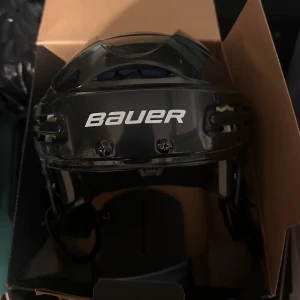 Svart hockeyhjälm Bauer BHH5100M - Säljer en svart hockeyhjälm från Bauer, modell BHH5100M. Hjälmen har vit logga framtill, justerbar passform och vadderad insida med blå och grå detaljer för extra komfort. Perfekt för dig som spelar hockey och vill ha en säker och snygg hjälm.