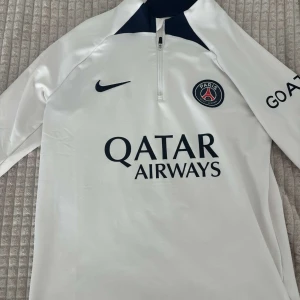 PSG långärmad träningströja Nike - Vit långärmad Paris Saint-Germain träningströja från Nike med mörkblå detaljer. Tröjan köptes från psg arenans officiella butik.