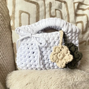 Handvirkad vit handväska med blommor - Superfin handvirkad vit handväska med tjockt garn och handtag. Väskan har en söt knytrosett och två virkade blommor i beige och olivgrönt som dekoration. Perfekt accessoar för att lyfta din outfit.