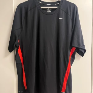 Nike Dri-Fit svart tränings-tshirt XL - Svart Nike Dri-Fit t-shirt i funktionsmaterial med röda detaljer längs sidorna. Klassisk rund halsringning och korta ärmar. Perfekt för löpning eller gym, med diskret Nike-logga på bröstet och ventilerande tyg.