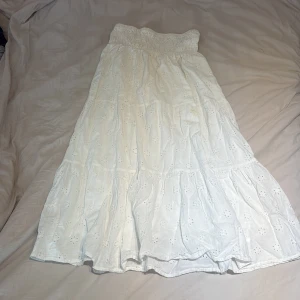 Vit långkjol med broderade blommor - Superfin vit långkjol med broderade blommor och smock i midjan. Kjolen är luftig och har en romantisk känsla med volanger och broderade detaljer. Perfekt för varma dagar och ger en bohemisk vibe till din outfit.