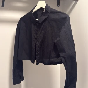 Svart croppad skjorta från Divided - Svart croppad skjorta från Divided med klassisk krage och knappar framtill. Skjortan har långa ärmar och en loose passform som ger en avslappnad vibe. Perfekt att styla med höga jeans eller kjol.