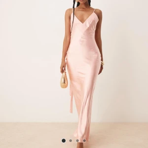 Ljusrosa långklänning i satin - Supersnygg ljusrosa långklänning i satin med smala axelband och v-ringning. Klänningen har en elegant rosettdetalj i ryggen och en figurnära passform som ger en lyxig känsla. Perfekt för dig som vill sticka ut med en stilren och trendig look.