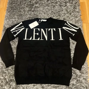 Svart stickad tröja från Valentino - Snygg svart stickad tröja från Valentino med stor vit logotyp över bröstet och ärmarna. Tröjan har rund halsringning, ribbade muddar och lång ärm. Perfekt för dig som vill ha en statement piece i garderoben.