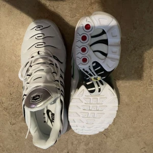 Nike Air Max Plus vita sneakers - Nike Air Max Plus sneakers i vitt med svarta detaljer och gröna accenter på sidan. Skorna har en unik vågig design, synlig Air Max-dämpning i sulan och silvrig detalj på plösen. Perfekta för dig som gillar streetwear och vill sticka ut med en modern look.