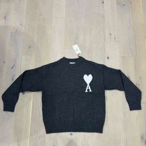 Mörkgrå stickad tröja från Ami Paris - Mörkgrå stickad tröja från Ami Paris med vit hjärtlogga och bokstaven A på bröstet. Tröjan har rund hals, ribbade muddar och är långärmad. Perfekt för dig som gillar stilren design med en twist.