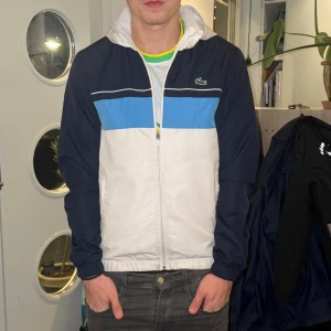 Lacoste vindjacka i blått och vitt - Vindjacka från Lacoste. Säljer pga att den inte kommer till andvändning. Den är i bra skick och har inte andvänds mycket. Köpt i tracksuit för ca 2800 kr. Pris kan diskuteras vid snabb affär