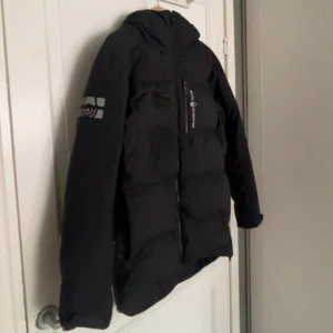 Svart dunjacka Sail Racing - Svart dunjacka från Sail Racing med huva och logga på bröstet samt ärmen. Jackan har dragkedja framtill, diskreta reflexdetaljer och en längre passform baktill. Perfekt för kalla dagar då den är extremt varm och är det perfekta exemplaret för just vintern, när du vill hålla stilen och värmen. Jackan är köpt för 3800 kr däremot går priset absolut att diskutera. 