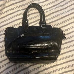 Svart handväska från Feel Accessories - Stilren svart handväska i glansigt skinn från Feel Accessories. Väskan har dubbla handtag, avtagbar axelrem och flera fack med dragkedja. Snygga silverdetaljer och klassisk fyrkantig form gör den enkel att matcha till olika outfits.