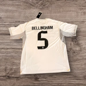 Real Madrid Bellingham tröja Adidas L - Säljer en officiell Real Madrid fotbollströja från Adidas med Bellingham och nummer 5 på ryggen. Tröjan är vit med svarta detaljer, gula inslag och klubbmärke på bröstet. Tillverkad i lätt funktionsmaterial (AEROREADY) och har klassiska svarta ränder på axlarna.