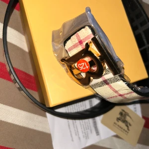 Burberry rutigt bälte med guldspänne - Snyggt bälte från Burberry med klassiskt rutigt mönster i beige, svart, rött och vitt. Bältet har ett stort guldigt B-spänne framtill och insidan är svart med präglad Burberry-logga. Perfekt accessoar för att lyfta din outfit.