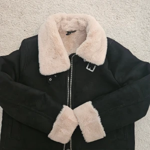 Svart teddyjacka från H&M Divided - Säljer en svart teddyjacka från H&M Divided i storlek M. Jackan har fluffigt beige foder och krage, dragkedja framtill och snygga spänndetaljer vid halsen. Perfekt för dig som vill ha en varm och trendig look under höst och vinter.