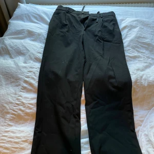 Svarta kostymbyxor i polyester - Svarta kostymbyxor med klassisk rak passform och bälteshällor. Tillverkade i en mjuk och skön blandning av polyester, viskos och elastan. Perfekta för en clean och stilren look. Midjan är normal och byxorna har diskreta fickor.