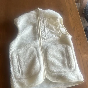 Vit fleeceväst med dragkedja från Studio Total - Mysig vit fleeceväst med hel dragkedja framtill och två stora fickor. Västen har en hög krage och är tillverkad i fluffigt syntetmaterial som ger en skön och avslappnad look. Perfekt att slänga på sig över en hoodie eller tröja.