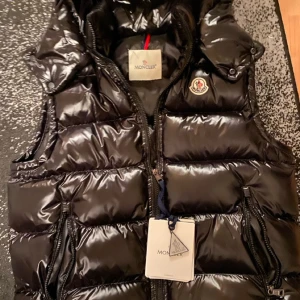 Moncler väst - Snygg svart dunväst från Moncler med glansig finish och stor vadderad huva. Västen har dragkedja framtill, två fickor med dragkedja och klassisk Moncler-logga på bröstet. Perfekt plagg för att dra blickar!                                                                Priset kan diskuteras vid snabb affär!