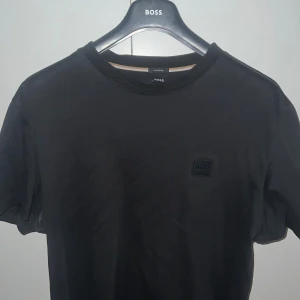 Svart t-shirt från BOSS med logga - Svart t-shirt från BOSS med klassisk rund hals och diskret svart logotyp-patch på bröstet. T-shirten är enkel och stilren, perfekt för dig som gillar minimalistisk design. Tillverkad i mjuk bomull för skön känsla hela dagen.