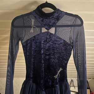 Mörkblå sammet konståknings klänning - Supersnygg mörkblå skridskoklänning i sammet och mesh med långärmade ärmar. Klänningen har glittriga detaljer och en cut-out vid halsen. Kjolen är kort och volangig. Svårt med storlekar men skulle säga den passar 152-158, jag är 158cm och passar ganska bra, lite kort i ärmarna!