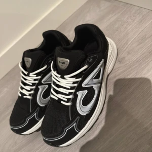Svarta sneakers med silvriga detaljer - Säljer ett par svarta sneakers med silvriga detaljer och vit sula. Skorna har vit snörning, mesh och syntetmaterial samt coola CD-loggor bak. Perfekta för dig som vill ha en lyxig och trendig sneaker med sportig vibe.