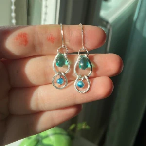 Silvriga örhängen med blå pärlor - Unika örhängen i silverfärgad metall med droppformad blågrön glasdetalj och liten blå pärla. Hängande modell med snyggt handgjord känsla och coola former. Perfekt för dig som gillar färg och vill sticka ut lite extra.