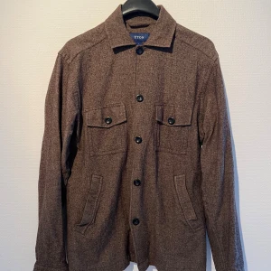 Brun overshirt från Eton i flanell - Snygg brun overshirt från Eton med fyra fickor och svarta knappar framtill. Jackan är gjord i mjuk flanell med bomull, ull och kashmir för extra komfort. Perfekt för lager-på-lager och har en klassisk krage och långärmad design.