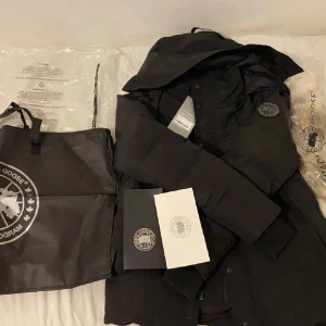 Svart dunjacka från Canada Goose L - Säljer en svart dunjacka  Canada Goose wyndham i storlek L. Jackan har huva, diskret logga på ärmen och stängs med både dragkedja och knappar. Klassisk pufferstil med quiltade sektioner och en clean look. Perfekt för kalla dagar. har även faktura och mer bilder på passformen