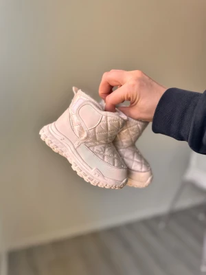 Beige quiltade vinterboots med foder för barn - Säljer ett par beige vinterboots med quiltad ovandel och mysigt foder inuti. Skorna har grov sula för bra grepp och stängs med kardborreband. Perfekta för kalla dagar och riktigt snygga till vinterlooken.