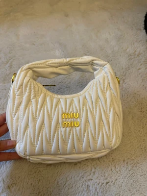 Vit quiltad handväska  - Supersnygg vit handväska med quiltad design och guldiga detaljer. Väskan har ett mjukt, vadderat handtag och guldfärgad logotyp framtill. Perfekt storlek för det viktigaste och ger en lyxig vibe till din outfit.