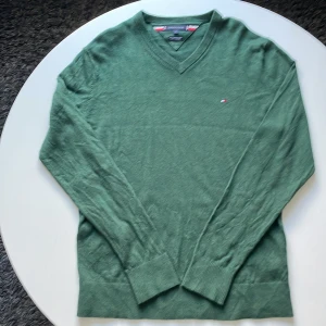 Grön v-ringad tröja Tommy Hilfiger - Snygg grön v-ringad tröja från Tommy Hilfiger i storlek L. Tillverkad i en mjuk blandning av pima bomull och kashmir. Diskret Hilfiger-logo broderad på bröstet. Långärmad med ribbade muddar och nederkant för extra komfort.