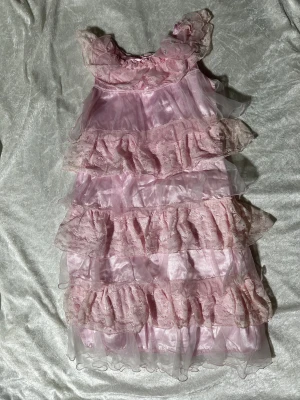 Barn Rosa volangklänning med spets och pärlor - Drömmig rosa långklänning med lager av volanger i spets och transparent tyg. Klänningen har dekorativa pärlor och en stor rosett vid halsen. Materialet är glansigt och ger en lyxig känsla, perfekt för dig som gillar romantiska detaljer och mjuka färger. Helt ny 