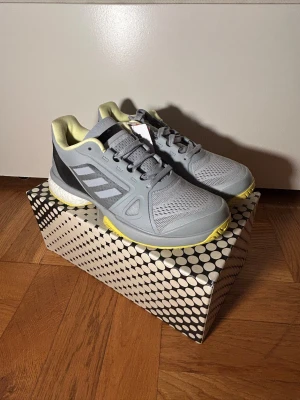 Adidas Stella McCartney grå sneakers - Snygga grå sneakers från Adidas Stella McCartney med ljusgula detaljer och vit boost-sula. Skorna har meshpaneler för extra ventilation, snörning och klassiska Adidas-ränder på sidan. Perfekt för dig som gillar sportig och modern stil. Extra sko snöre. (Ordinare pris 1400kr)