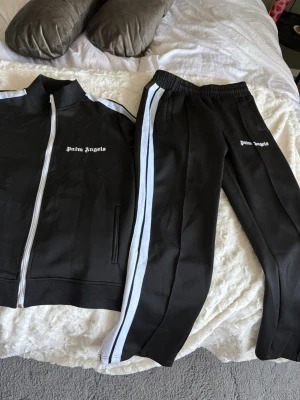 Palm Angels mjukisbyxor - Svarta mjukisbyxor från Palm Angels med vita sidoremsor och logga tryckt på benet. Byxorna har elastisk midja och raka ben, tillverkade i ett glansigt syntetmaterial som ger en sportig vibe. Kan tänka mig att sälja byxorna och tröjan separat! 800 för byxor, 600 för tröjan