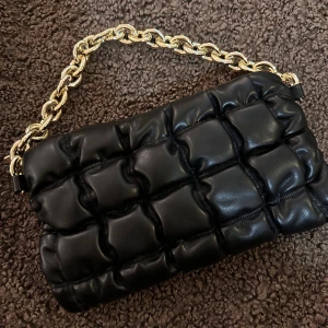 Svart quiltad axelväska med kedja - Snygg svart axelväska med quiltad design och en chunky guldfärgad kedja som handtag. Väskan är gjord i skinnimitation och har dragkedja upptill samt flera fack inuti. Perfekt storlek för det viktigaste och ger en cool vibe till din outfit.