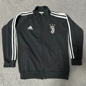 Juventus svart Adidas track jacket - Svart Adidas track jacket med Juventus-logga och tre guldstjärnor broderat på bröstet. Klassiska vita ränder längs ärmarna, hel dragkedja och ribbade muddar. Tillverkad i polyester och perfekt för fotboll eller streetwear.