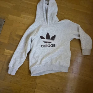 Grå Adidas hoodie med huva - Snygg grå hoodie från Adidas med det klassiska treklöver-loggan i svart på bröstet. Tröjan har huva och långa ärmar med ribbade muddar. Perfekt för chill dagar eller när du vill ha en sportig look.