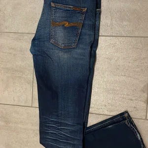 Mörkblå Nudie Jeans, storlek  - Snygga mörkblå jeans från Nudie Jeans med klassisk orange broderad våg på bakfickorna. Jeansen har en straight passform och är tillverkade i slitstarkt denim med coola slitningar och kontrastsömmar. Perfekt för dig som gillar street och casual stil. 33/34