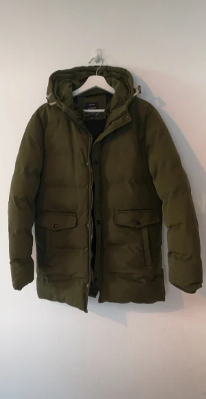 Olivgrön pufferjacka - Olivgrön pufferjacka från Jing Pin Men med huva och snörning. Jackan har två stora fickor framtill med lock och knappar, samt dragkedja och knäppning framtill. Jackan har aldrig blivit använd.