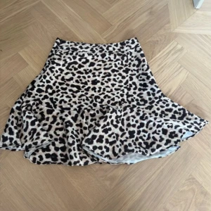Leopardmönstrad volangkjol från Amisu - Snygg kort kjol från Amisu med leopardmönster i beige och svart. Kjolen har volang nertill som ger en cool och trendig look. Perfekt för dig som vill sticka ut med ett djurmönster och gillar att leka med stilen.