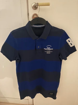 Blå randig Peak Performance piké - Snygg pikétröja från Peak Performance i marinblått och mörkblått randigt mönster. Klassisk krage, korta ärmar och broderad vit 86 på ena ärmen. Materialet är mjuk bomull och tröjan har knappar framtill. Perfekt för dig som gillar sportig stil.