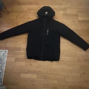 Svart vindjacka från Sail Racing - Svart vindjacka från Sail Racing med huva och dragkedja framtill. Jackan är vattentät, vindtät och har andningsfunktion. Primaloft-foder. Snygg logga på bröstet och huvan, samt praktiska fickor och justerbara ärmslut. Modellen är 182 och den sitter lite kort i båda armarna och midjan. Priset går att dikutera 