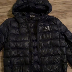 Mörkblå EA7 dunjacka med huva - Snygg mörkblå dunjacka från EA7 Emporio Armani med huva och tydlig logga på bröstet. Jackan är quiltad och har dragkedja med EA7-detalj. Perfekt för dig som vill ha en stilren och varm jacka med exklusiv känsla.